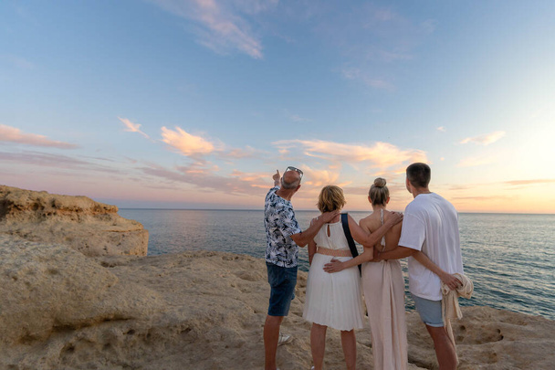 夏の旅行中に海の上の絵のような日没を賞賛する岩の崖の上に立っている家族と一緒に空を指す男の背景ビュー - 写真・画像