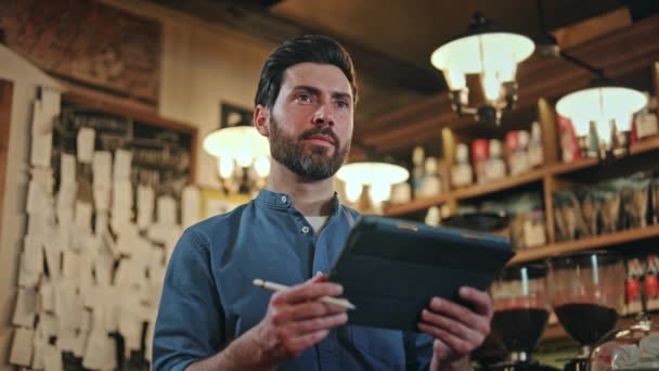 Přátelský běloch v modré uniformě přidává informace do digitálního tabletu během pracovního procesu v baru. Kompetentní zaměstnanec s perem stylus v ruce s objednávkou od hosta na moderním pracovišti. - Záběry, video