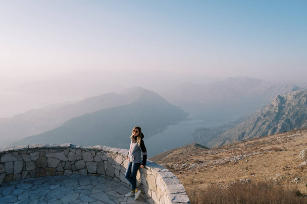 Chica con gafas de sol en una chaqueta se encuentra junto a una valla de piedra en las montañas por encima de una bahía en la niebla. Foto de alta calidad - Foto, imagen