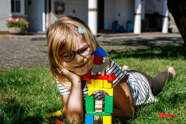 屋外のテーブルの上の木製のブロックで遊んでいる小さな幼稚園の少女. 小さな子供のための教育ゲーム. 幸せな子供は木製の虹スタッキングブロックからタワーを構築します - 写真・画像