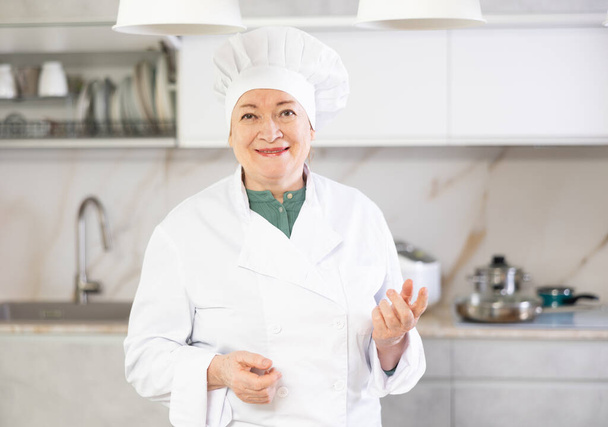 Happy mature female cook in uniform standing and showing her kitchen - Foto, Bild