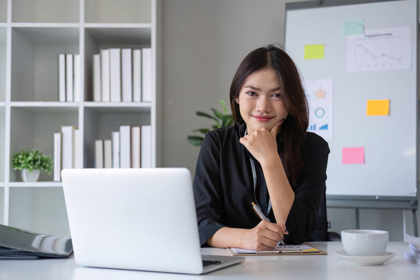Красивая улыбающаяся азиатская бизнесвумен, работающая на ноутбуке и пьющая кофе азиатская бизнесвумен, работающая с финансами и калькулятором в своем офисе. - Фото, изображение