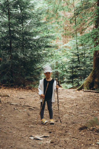 Malá dívka cestovatel stojí na skalnaté horské stezce mezi vysokým jehličnatým lesem s holí v rukou. - Fotografie, Obrázek
