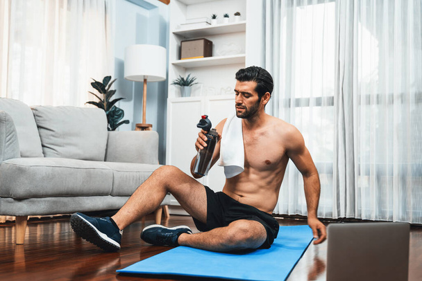Athletic and sporty man drinking water on fitness mat after finishing home body workout exercise session for fit physique and healthy sport lifestyle at home. Gaiety home exercise workout training. - Photo, Image
