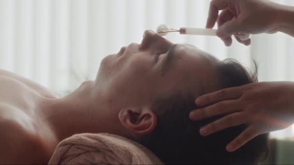 Acercamiento lateral de esteticista haciendo masaje facial a cliente masculino usando rodillo de jade en estudio de spa - Metraje, vídeo