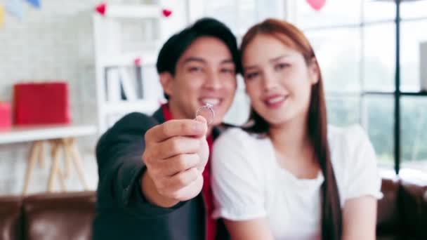 Close-up hand of the man holding the ring in front of the camera with a blurred couple in love sitting together in the background. Valentine's Day celebration concept. Wedding and engagement concept. - Footage, Video