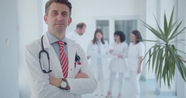 Portret van een zelfverzekerde arts in een kliniek, staande met gevouwen armen. Gelukkig en professioneel, omringd door andere dokters achterin. - Video