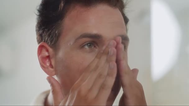 Groot close-up van gen Z man in witte badjas ribbing voedende crème in het gezicht in de voorkant van de spiegel, het verzorgen van de huid - Video