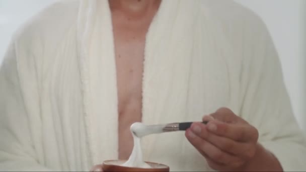 Tilt up shot του GEN Z άνθρωπος σε λευκό μπουρνούζι ανάμειξη και στη συνέχεια την εφαρμογή κρέμα ξυρίσματος στο πρόσωπο με πινέλο μπροστά από τον καθρέφτη στο σπίτι - Πλάνα, βίντεο