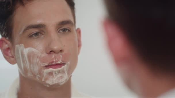 Primer plano del hombre afeitándose la cara con una navaja mientras se prepara para el trabajo por la mañana de pie frente al espejo - Imágenes, Vídeo