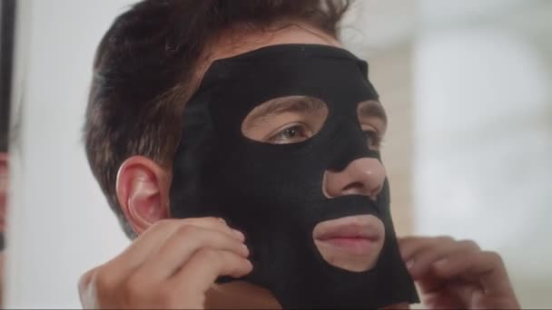 Μεγάλο κοντινό πλάνο του νεαρού με μικτή εθνικότητα χρησιμοποιώντας μαύρη ενυδατική μάσκα στο πρόσωπο κατά τη διάρκεια των διαδικασιών φροντίδας του δέρματος μπροστά από τον καθρέφτη στο μπάνιο - Πλάνα, βίντεο