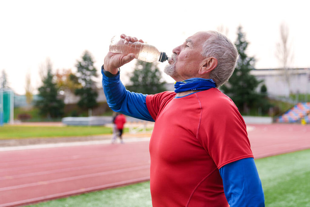 Atleta maduro bebendo água de uma garrafa, fazendo uma pausa de hidratação em uma pista de esportes - Foto, Imagem