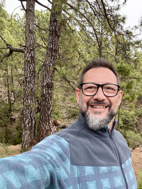 Счастливый весельчак один в лесу фотографирует селфи на свежем воздухе, развлекаясь в одиночестве с деревьями и лесом на заднем плане. Современный путешественник наслаждается живописным местом на горе. Природа - Фото, изображение