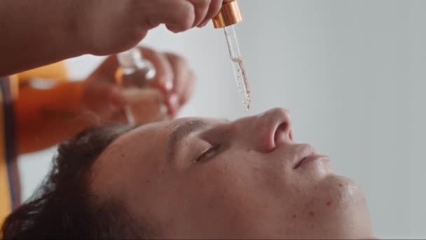 Naklonění ruky kosmetičky aplikující hydratační olej na tvář mužského klienta během lázeňské procedury v salonu - Záběry, video