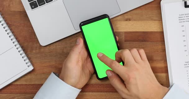 Чоловік переміщує індексний палець на зелений порожній екран сучасного смартфона на робочому місці зверху. Крупним планом руки людини тримає мобільний телефон у вертикальному положенні - Кадри, відео