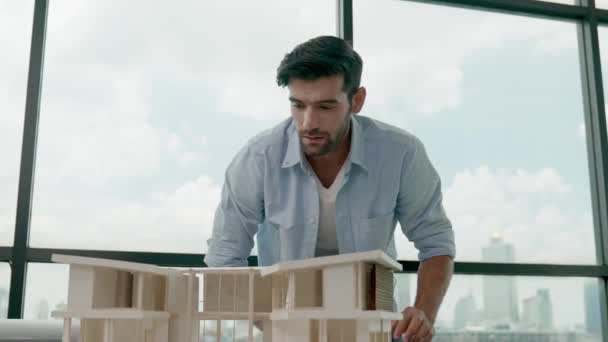 Έξυπνος πολιτικός μηχανικός μετράει το μοντέλο του σπιτιού και γράφει στο σχεδιάγραμμα. Επαγγελματίας μηχανικός μέτρο, μοντέλο σπίτι σχεδιασμού, ενώ στέκεται κοντά παράθυρο πανόραμα με ουρανοξύστη. Ίχνη - Πλάνα, βίντεο