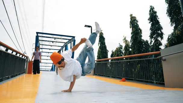 Panorama shot van professionele hipster uit te voeren b-boy prestaties en energieke voetstap. Bekwame gelukkige man oefent breakdance terwijl een vriend hem aanmoedigt op de brug. Outdoor sport 2024. Schitterend.. - Foto, afbeelding