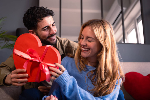 Όμορφο νεαρό ζευγάρι στο σπίτι απολαμβάνει το χρόνο μαζί, ενώ γιορτάζει την Ημέρα του Αγίου Βαλεντίνου με ένα κουτί δώρου σε σχήμα καρδιάς στο χέρι - Φωτογραφία, εικόνα