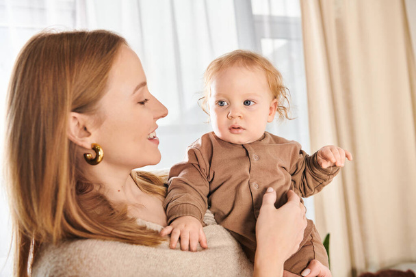 jeune femme souriante tenant le petit garçon dans les mains dans la chambre d'enfant à la maison, maternité heureuse - Photo, image