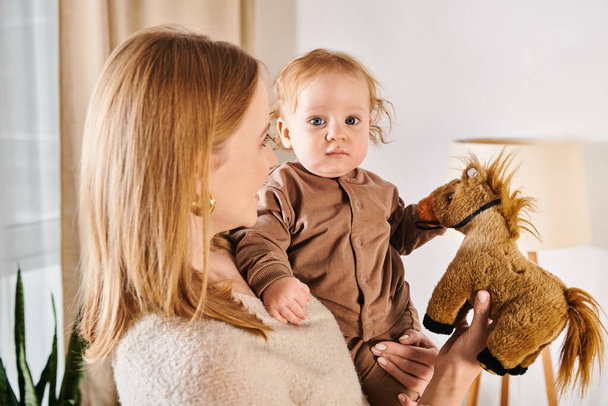 junge Frau hält niedlichen Sohn und Spielzeugpferd in den Händen im Kinderzimmer zu Hause, glückliche Mutterschaft - Foto, Bild