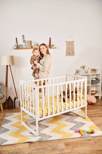 szczęśliwa młoda kobieta z uroczym synkiem w rękach stojących w pobliżu łóżeczka w pokoju dziecinnym, macierzyństwo - Zdjęcie, obraz