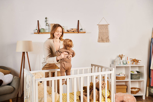 молодая веселая мать держит маленького ребенка сына возле кроватки в детской комнате, блаженное материнство - Фото, изображение
