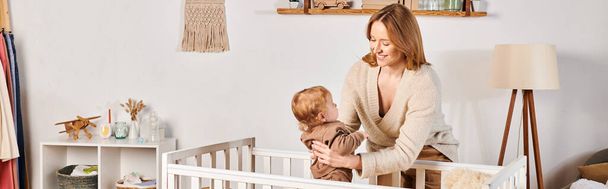 счастливая молодая мать держит милый малыш сын рядом с удобной кроваткой в детской комнате, горизонтальный баннер - Фото, изображение