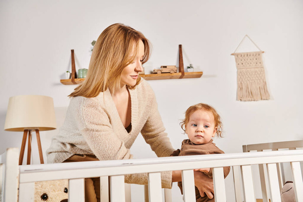 счастливая молодая мать, поддерживающая маленького ребенка, стоящего в кроватке в детской комнате дома, материнство - Фото, изображение