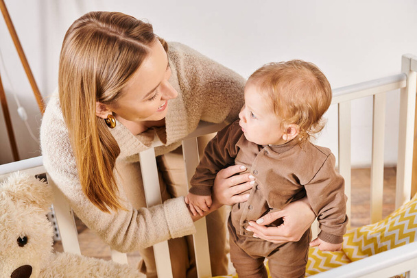 šťastná matka podporující rozkošný chlapeček stojící v postýlce v dětském pokoji doma, prapor - Fotografie, Obrázek