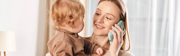 alegre madre multitarea sosteniendo al hijo en las manos y hablando en el teléfono inteligente en la habitación de la guardería, pancarta - Foto, Imagen