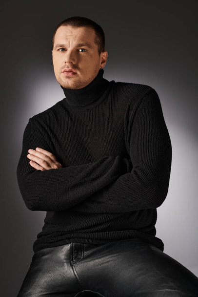 задумчивый харизматичный модный мужчина в черном свитере смотрит в камеру на сером фоне с освещением - Фото, изображение