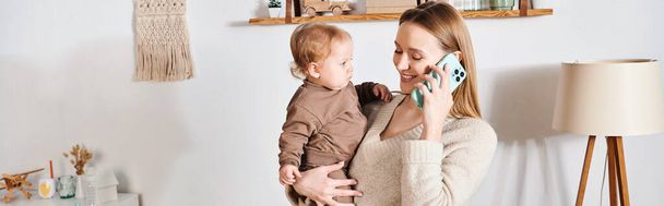 smiling woman with toddler son in hands talking on mobile phone in nursery room, horizontal banner - Photo, Image