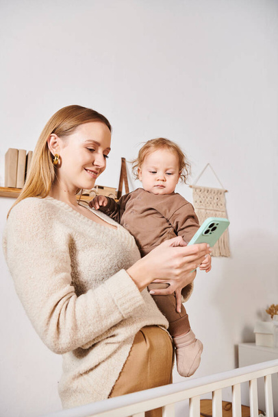 młoda matka z małym synkiem w ręku wiadomości przez telefon komórkowy w pokoju dziecinnym, wielozadaniowa kobieta - Zdjęcie, obraz