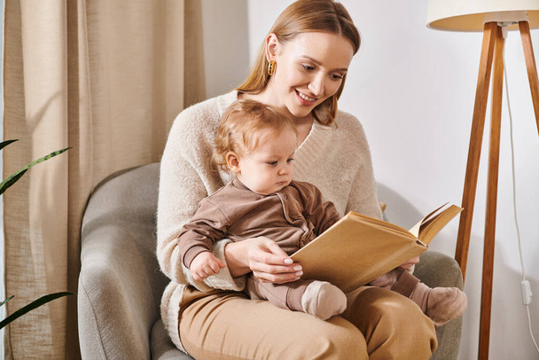glückliche Mutter liest Buch zu entzückenden Kleinkind Sohn im Sessel in gemütlichen Kinderzimmer, moderne Elternschaft - Foto, Bild