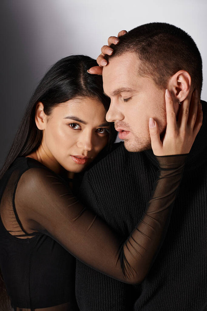 brunette passionate asian woman in black casual attire embracing head of stylish man on grey - 写真・画像