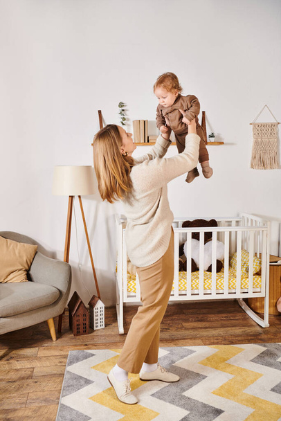 молода жінка тримає маленького сина, граючи і розважаючись біля ліжечка в дитячій кімнаті, материнство - Фото, зображення