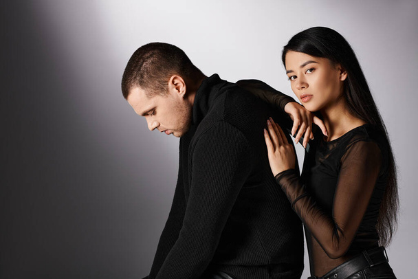 очаровательная азиатская женщина опираясь на мужчину в черной одежде и глядя на камеру на серый, любовь и моду - Фото, изображение