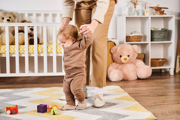 mother holding hands of toddler son walking near toys and crib in nursery room, care and support - Photo, Image