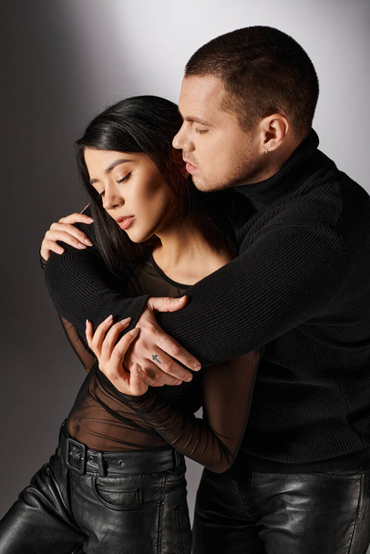 handsome man in black outfit embracing alluring asian woman on grey backdrop, love and fashion - Foto, Imagen