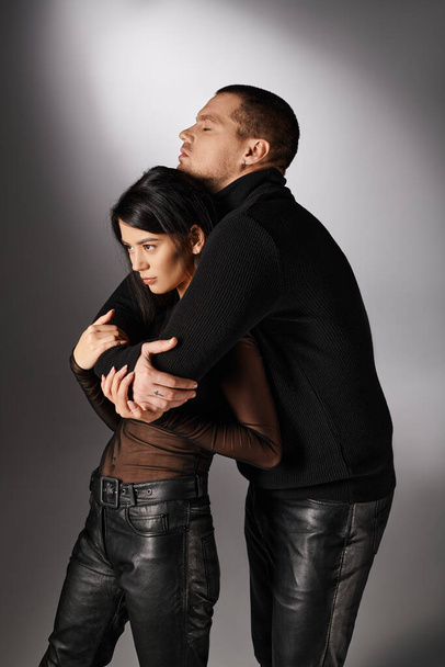 trendi karizmatikus férfi fekete ruhában átöleli ázsiai nő szürke háttér, szerelem és divat - Fotó, kép
