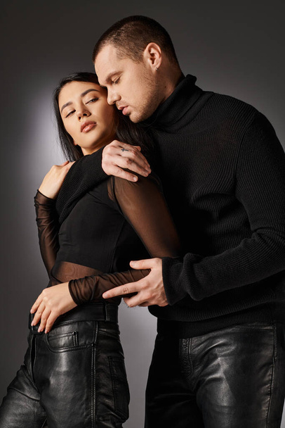 tender handsome man in black clothes hugging asian woman on grey backdrop, love and fashion - Photo, Image