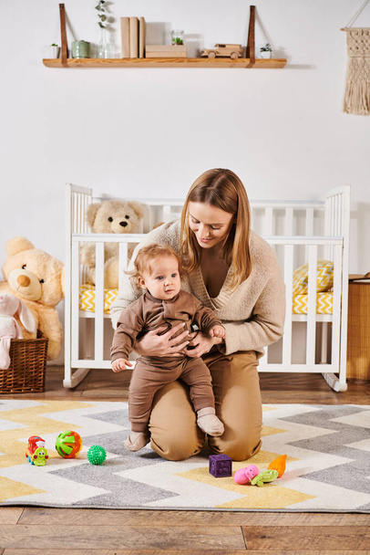 joyful woman embracing toddler boy near toys on floor in cozy nursery room, happy motherhood - 写真・画像
