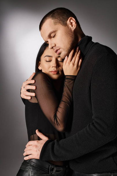 amante pareja interracial en negro ropa de moda abrazando con los ojos cerrados en gris telón de fondo - Foto, imagen