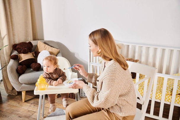 νεαρή γυναίκα σίτιση νήπιο γιο με πρωινό σε καρέκλα μωρού στο παιδικό δωμάτιο, σύγχρονη ανατροφή των παιδιών - Φωτογραφία, εικόνα