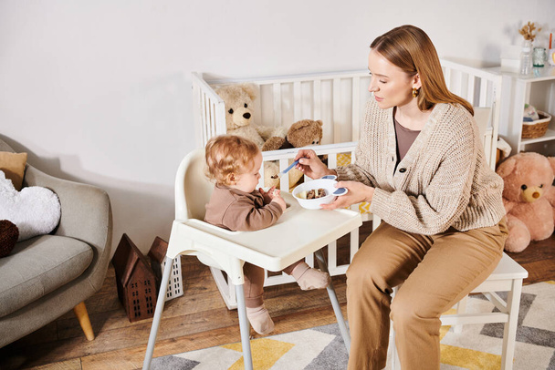 İlgili bir kadın, bebeği bebek koltuğunda kahvaltıyla besliyor. Bebek odasında, mutlu bir annelik. - Fotoğraf, Görsel