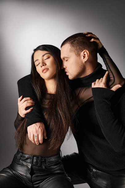 trendy interracial couple in black casual attire embracing with closed eyes on grey backdrop - Foto, Imagem
