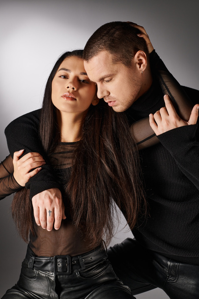 спокуслива азіатська жінка в чорному одязі дивиться на камеру біля молодого чоловіка, обіймаючи її на сірому - Фото, зображення