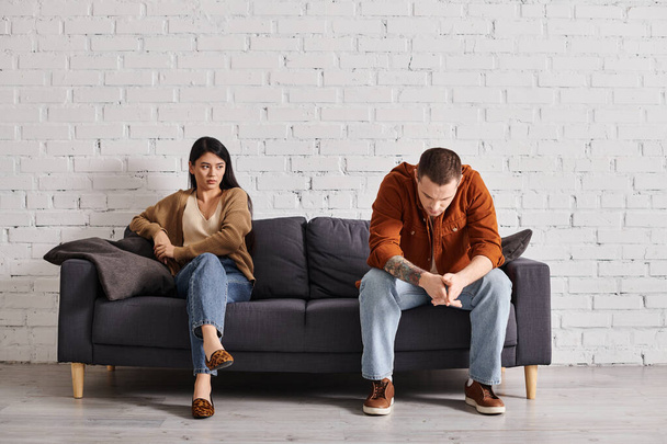 νεαρό απογοητευμένο διαφυλετικό ζευγάρι κάθεται στον καναπέ στο σαλόνι, έννοια του οικογενειακού διαζυγίου - Φωτογραφία, εικόνα