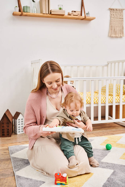 щаслива мати грає з милим маленьким сином біля ліжечка на підлозі в дитячій кімнаті, блаженне материнство - Фото, зображення