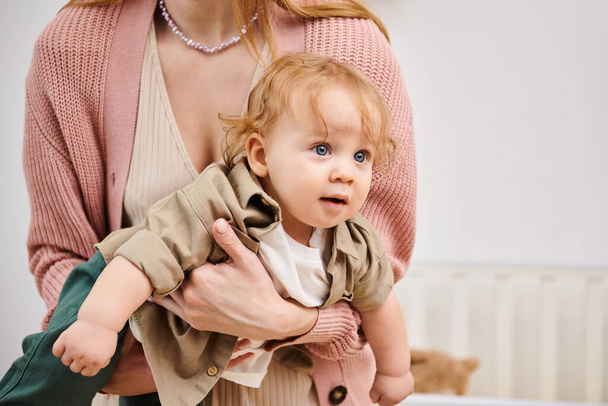 adorable bébé garçon regardant loin dans les mains de sa mère dans la chambre d'enfant à la maison, enfance heureuse - Photo, image